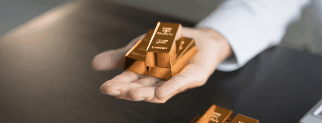 An outstretched hand holding or offering a stack of 3 small gold bars, with more gold bars on the desk beneath the arm.