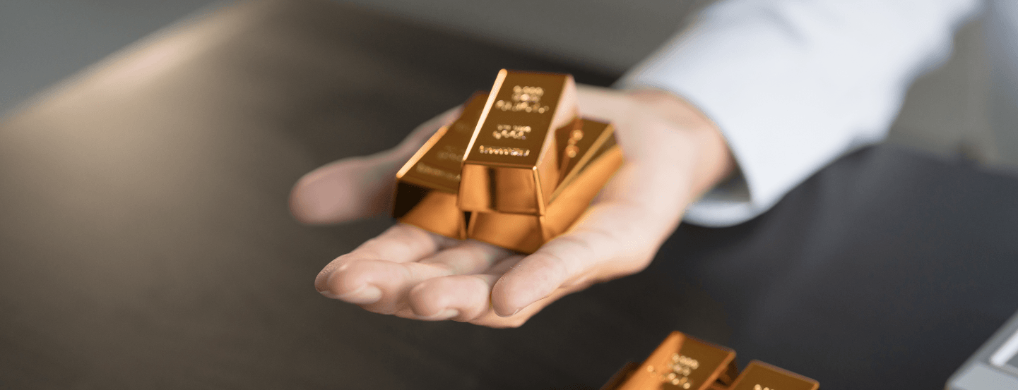 An outstretched hand holding or offering a stack of 3 small gold bars, with more gold bars on the desk beneath the arm.